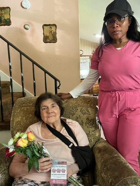 A caregiver in pink scrubs stands beside an older woman seated in a chair holding a bouquet of roses, sharing a caring moment in a cozy home.