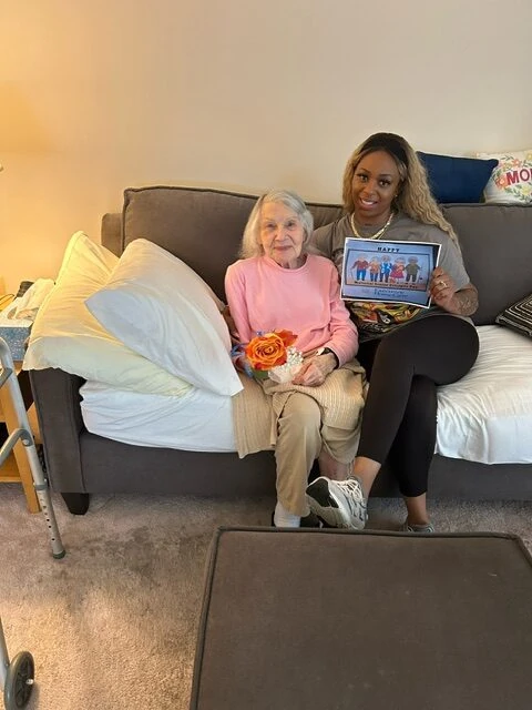 A caregiver sits beside an elderly woman on a couch, holding a sign and a rose while sharing a warm and celebratory moment.