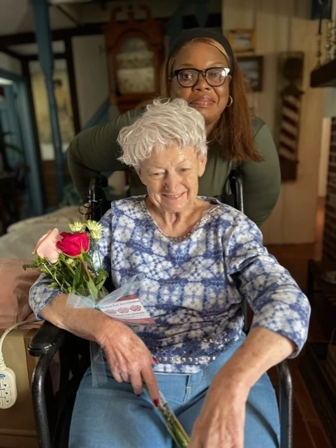 A caregiver stands behind an elderly woman seated in a wheelchair, both smiling as the woman holds a rose.