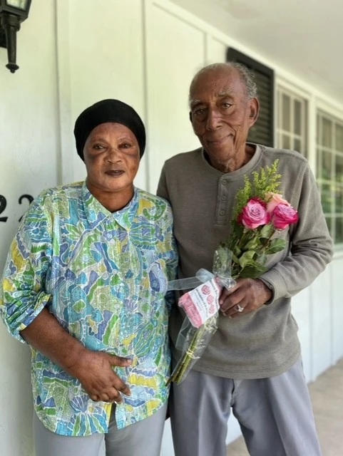 An older couple stands together on a porch holding a bouquet of flowers, sharing a quiet and meaningful moment.