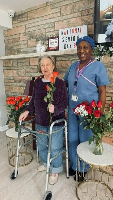 An older woman stands with a care giver holding a rose; both are smiling. A sign in the back reads "National Senior Day".