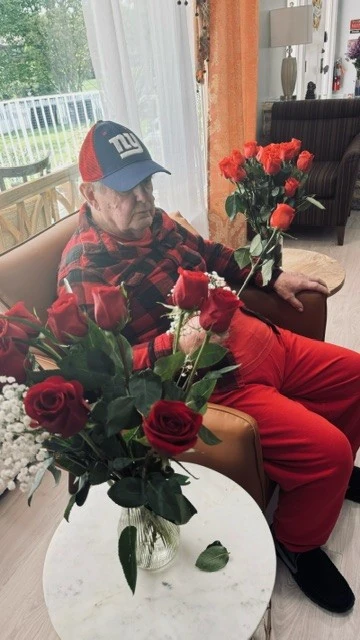 An older man sits in a chair holding a bouquet of red roses near a window, surrounded by soft natural light.