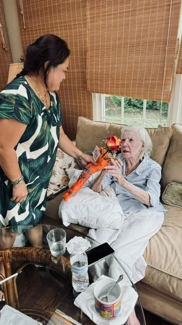 A care giver gifts a rose to an elderly woman sitting on a couch in front of a window in natural light.