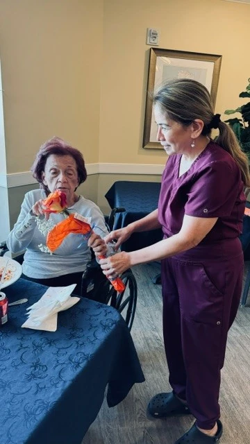 A care giver hands an elderly woman in a wheelchair a rose as they discuss the meaning of the rose.