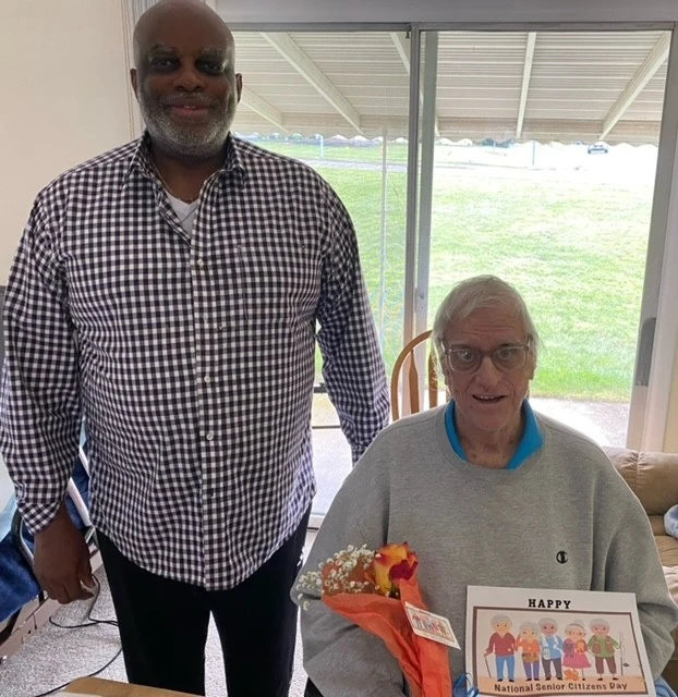 An older man sits holding a bouquet and a “Happy National Senior Citizens Day” sign while a caregiver stands beside him, sharing a proud and celebratory moment near a bright window.