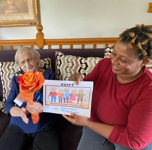 A caregiver sits beside an elderly woman on a couch, holding a celebratory sign while the woman holds a rose, both sharing a joyful and appreciative moment.