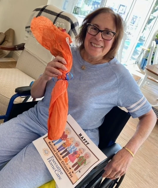 An older woman in a wheelchair smiles while holding a wrapped rose and a “Happy National Senior Citizens Day” sign in a bright indoor setting.