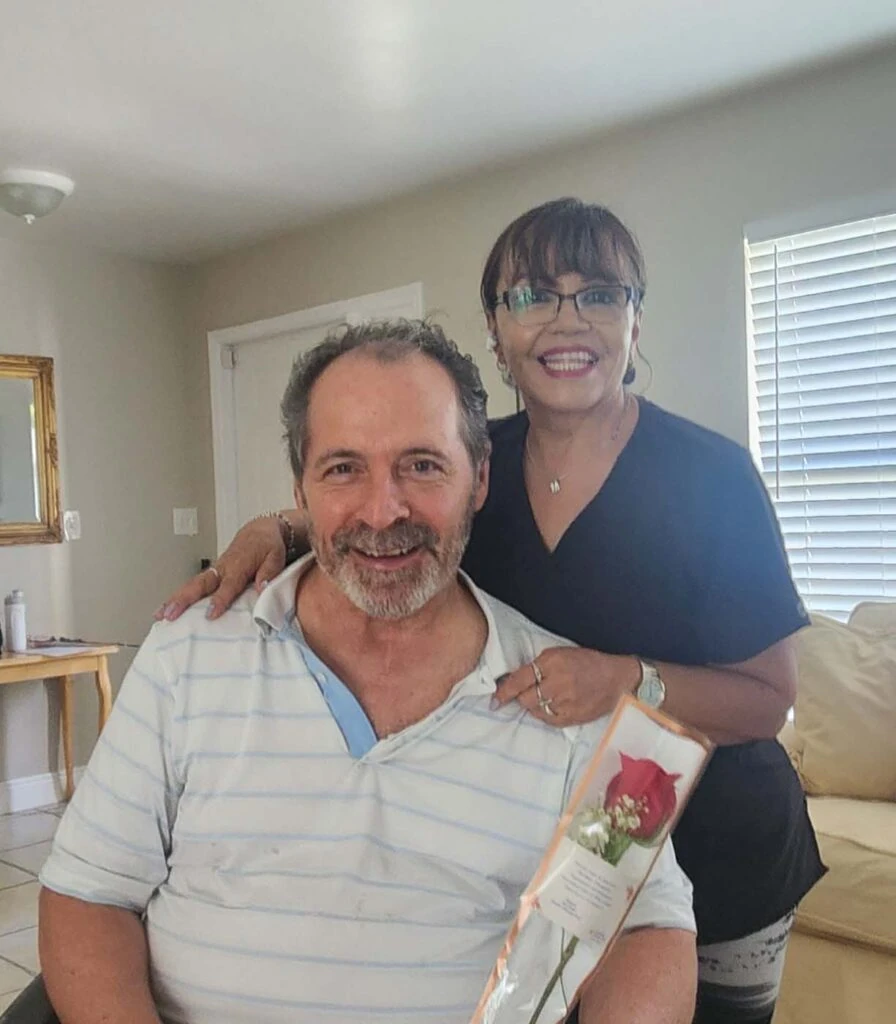 A smiling caregiver stands beside a seated man, resting a hand on his shoulder as he holds a wrapped rose, sharing a warm and appreciative moment in a home setting.
