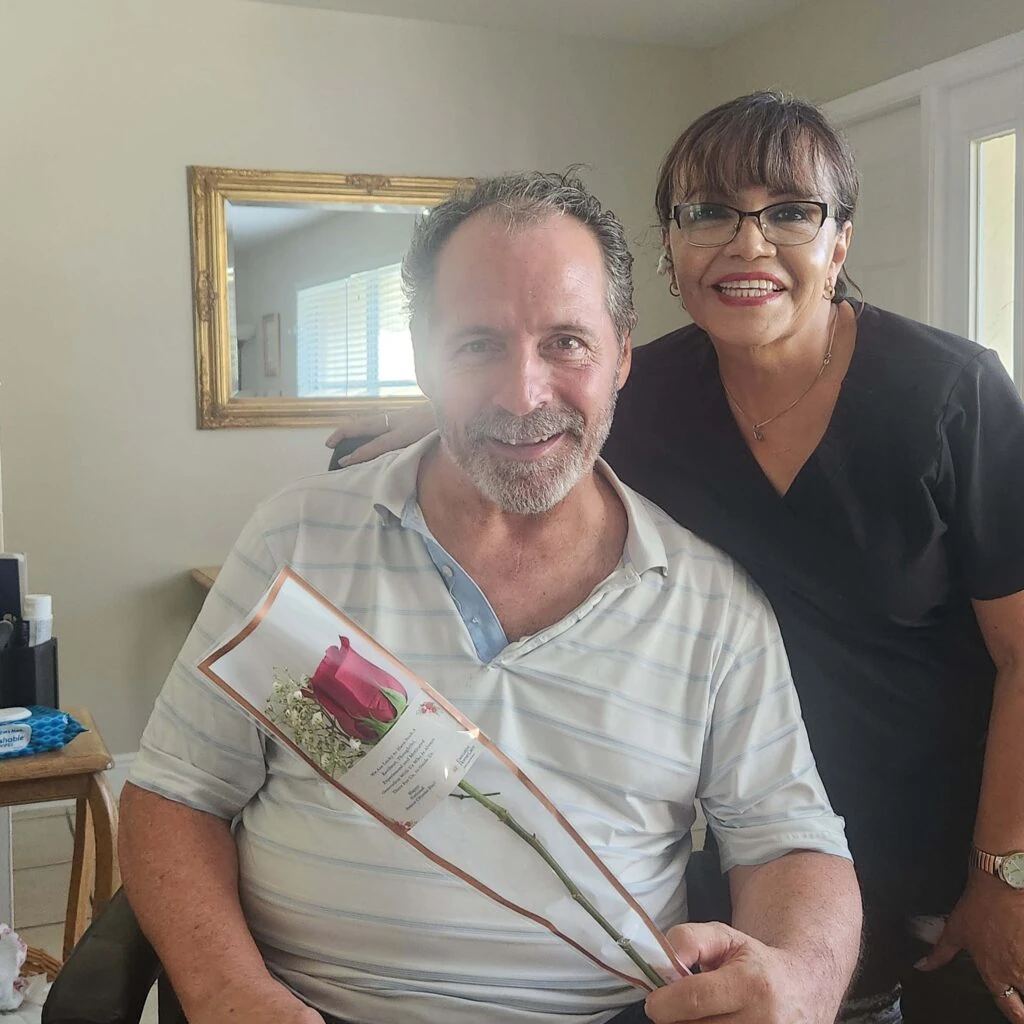 A smiling man sits holding a wrapped rose while a caregiver stands beside him with a hand on his shoulder, both posing together in a warm home environment.
