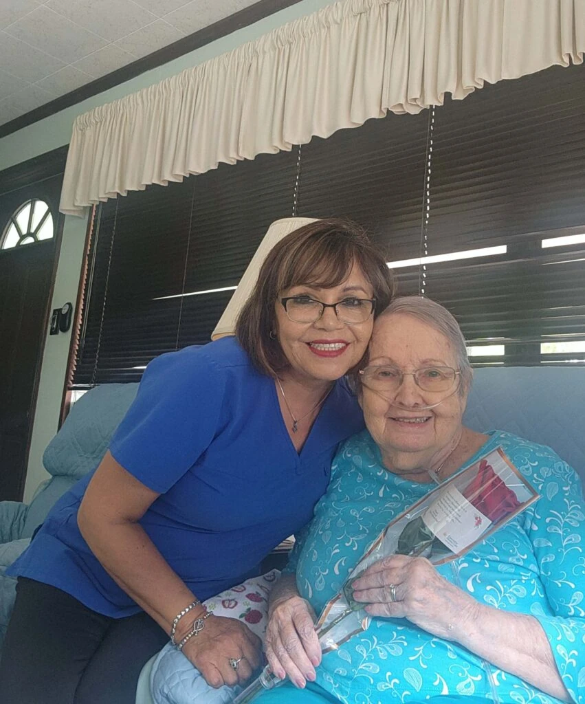 A caregiver leans in beside an elderly woman seated comfortably, both smiling as the woman holds a wrapped rose, sharing a warm and affectionate moment indoors.
