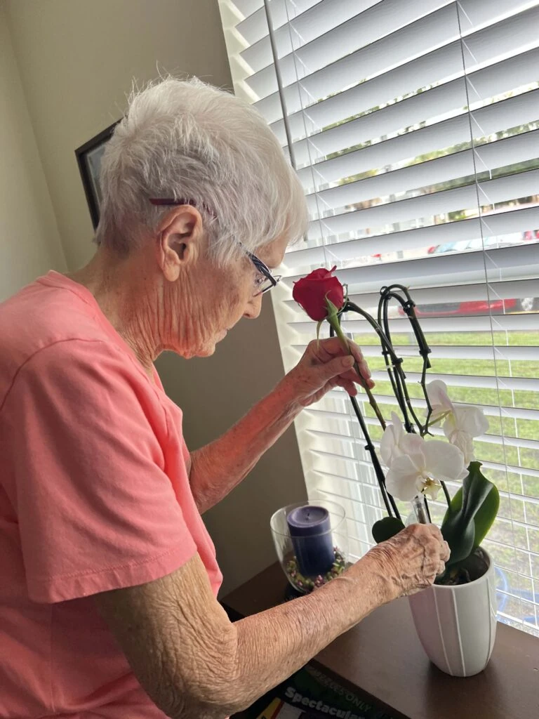An elderly woman stands by a window arranging a single red rose into a white orchid plant on a table, concentrating gently as daylight filters through the blinds.
