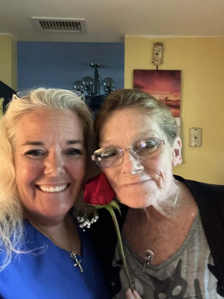 A caregiver and an older woman pose closely together indoors, both smiling warmly as the older woman holds a single red rose near her face.