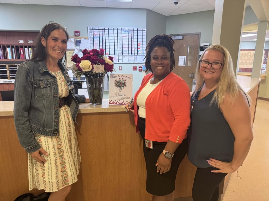 Three caregivers stand behind a reception desk smiling beside a vase filled with red and white roses and a sign promoting a “Roses for Resilience” initiative.