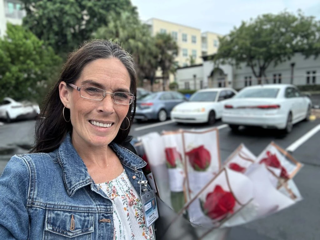 A smiling caregiver stands in a parking lot holding several wrapped red roses, with cars and a residential building in the background.