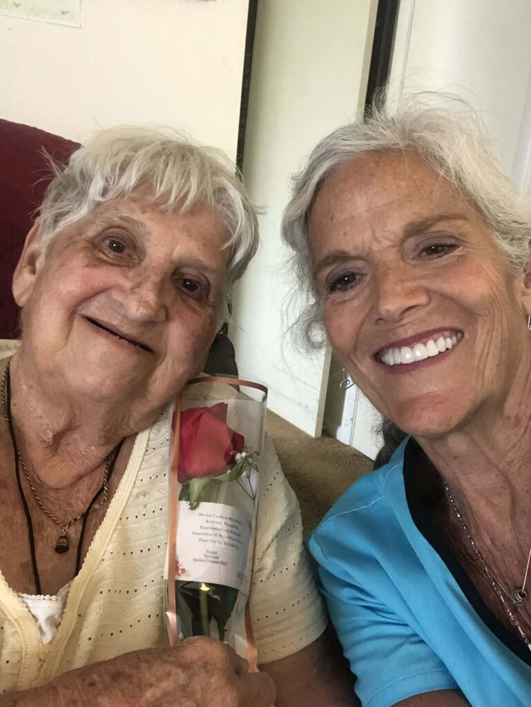 Two women smile together for a close-up selfie, with one holding a wrapped red rose, creating a warm and celebratory moment.