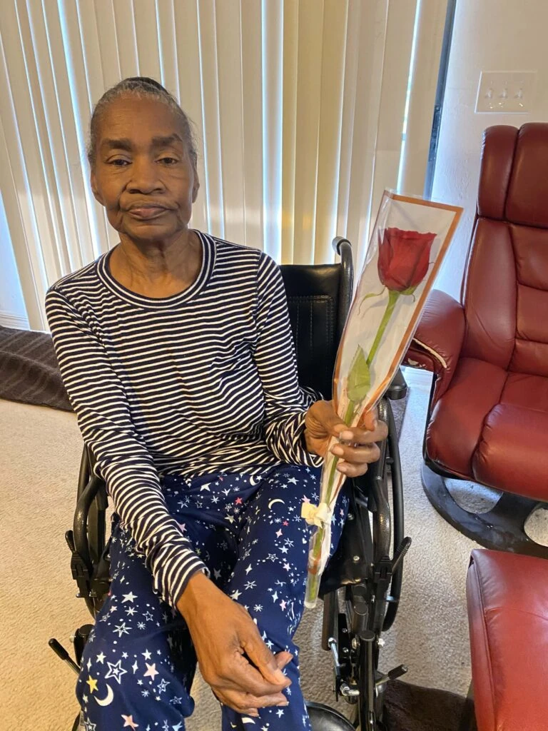 An elderly woman in a wheelchair holds a single wrapped red rose indoors, seated beside a red recliner chair and window blinds.