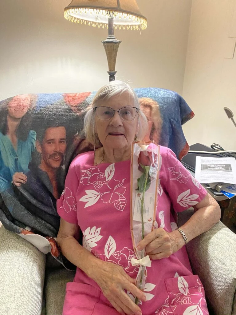An elderly woman in a pink floral dress sits in a cushioned chair holding a single wrapped red rose, with a lamp and blanket behind her in a cozy living room.