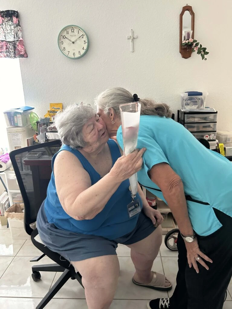 An older woman seated in a chair leans in to hug a caregiver while holding a wrapped red rose, capturing a warm and affectionate moment indoors.