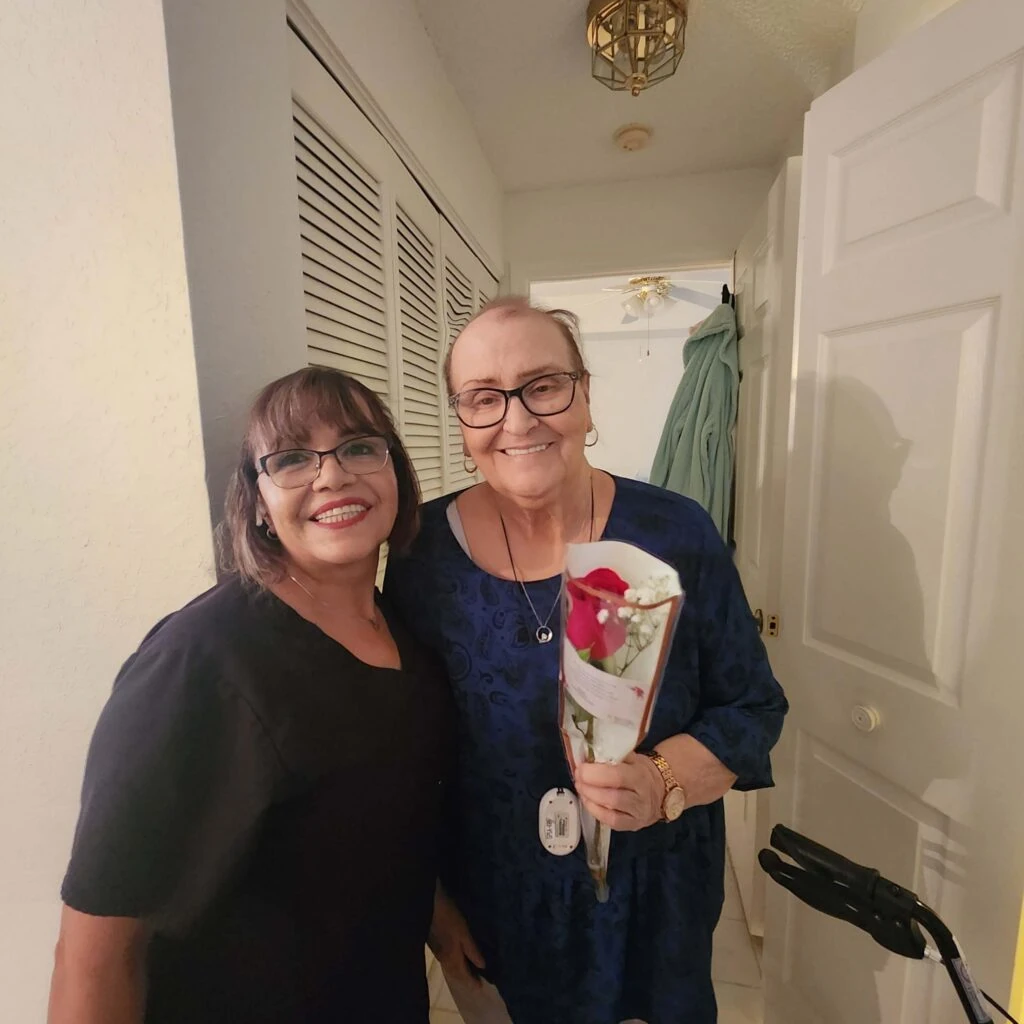 Two women stand together smiling in a hallway, one holding a wrapped red rose while a walker is visible nearby.