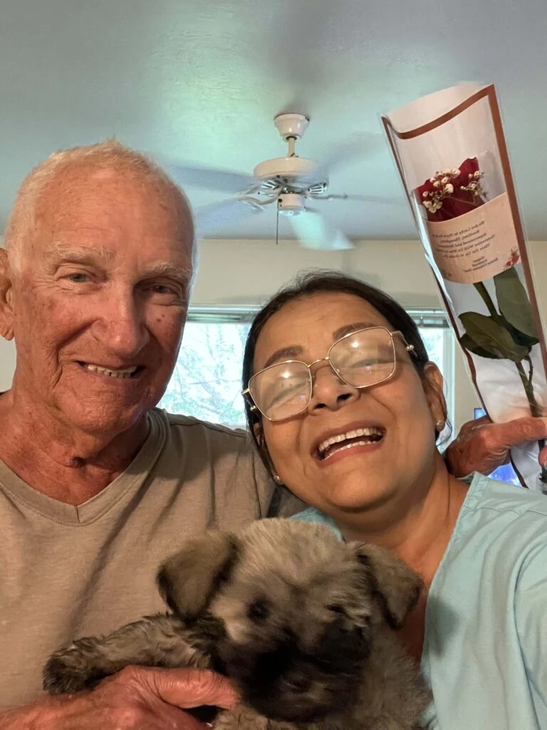 An older man and a caregiver smile together indoors while holding a small dog, with a wrapped red rose visible beside them.