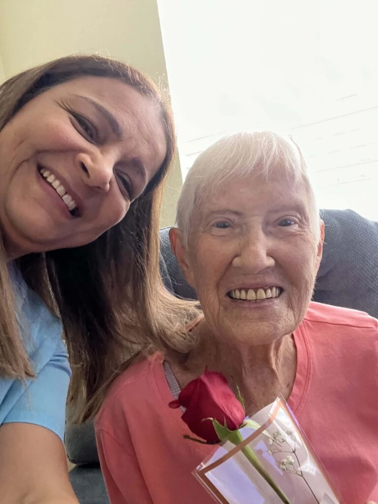 A caregiver leans in beside a smiling elderly woman holding a wrapped red rose, capturing a cheerful selfie moment indoors.