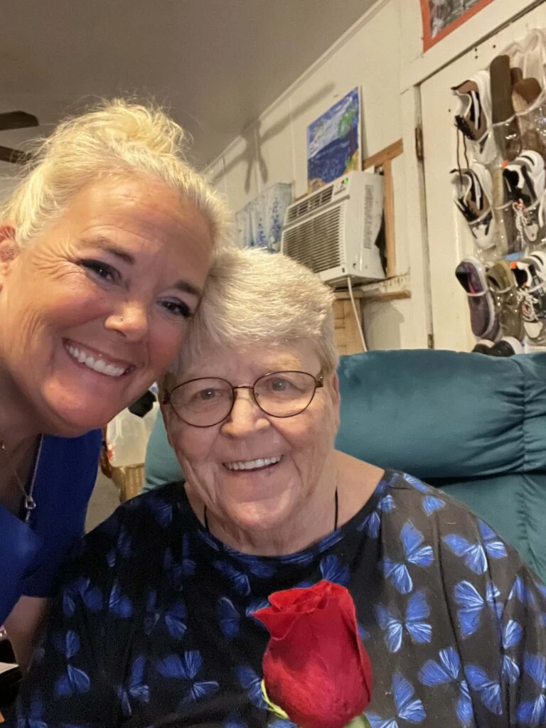 A caregiver and an elderly woman smile closely for a selfie, with the older woman holding a single red rose in a cozy home setting.