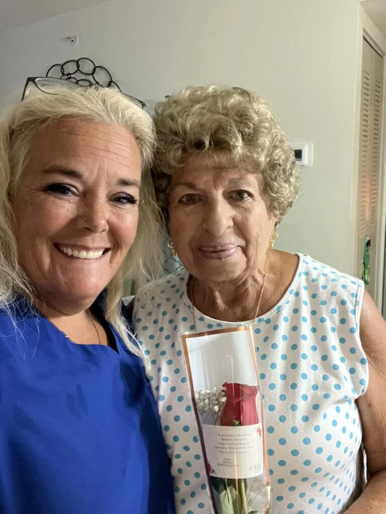 A caregiver and an elderly woman smile together for a photo, with the older woman holding a wrapped red rose.