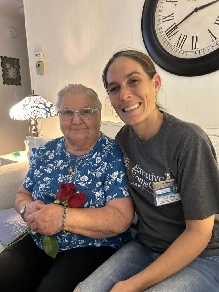 A caregiver sits beside an elderly woman on a couch, both smiling as the woman holds several red roses in a warmly lit living room.