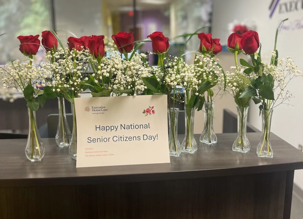 Multiple small vases of red roses with white filler flowers line a table beside a sign reading “Happy National Senior Citizens Day!”
