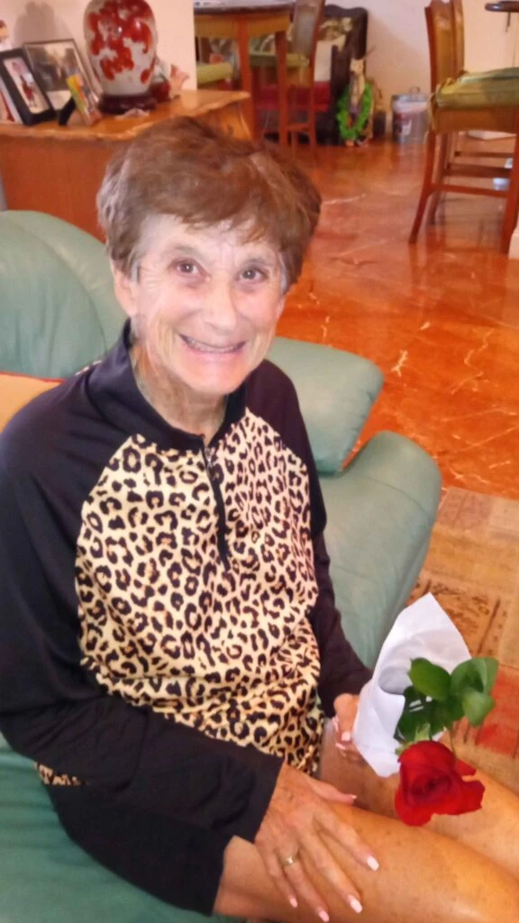 An elderly woman sitting on a couch smiles while holding a single red rose wrapped in paper.