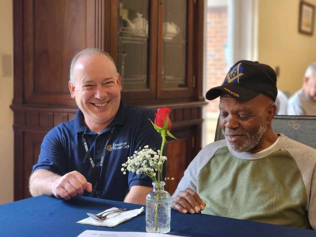 A caregiver and an elderly man sit together at a table, smiling as they look at a red rose arranged in a small glass vase.