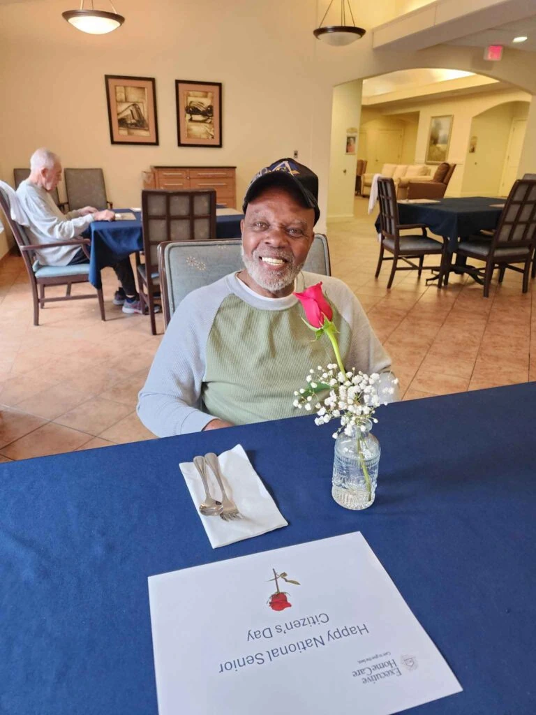 A smiling elderly man sits at a dining table with a red rose in a small vase, with a sign reading “Happy National Senior Citizens Day” in front of him.