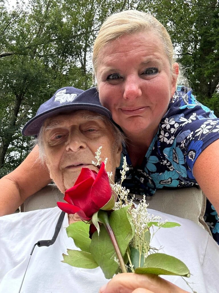 A caregiver leans in smiling beside an elderly man outdoors as he holds a single red rose.