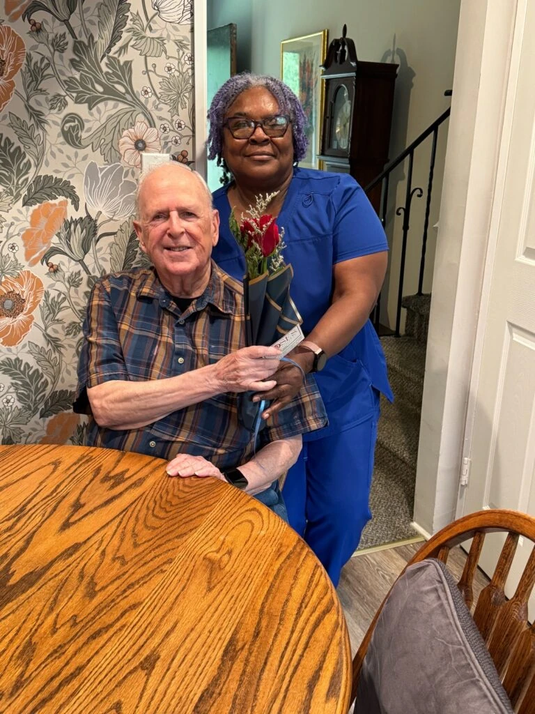 A caregiver in blue scrubs stands beside an elderly man seated at a table, both smiling as he holds a wrapped red rose.
