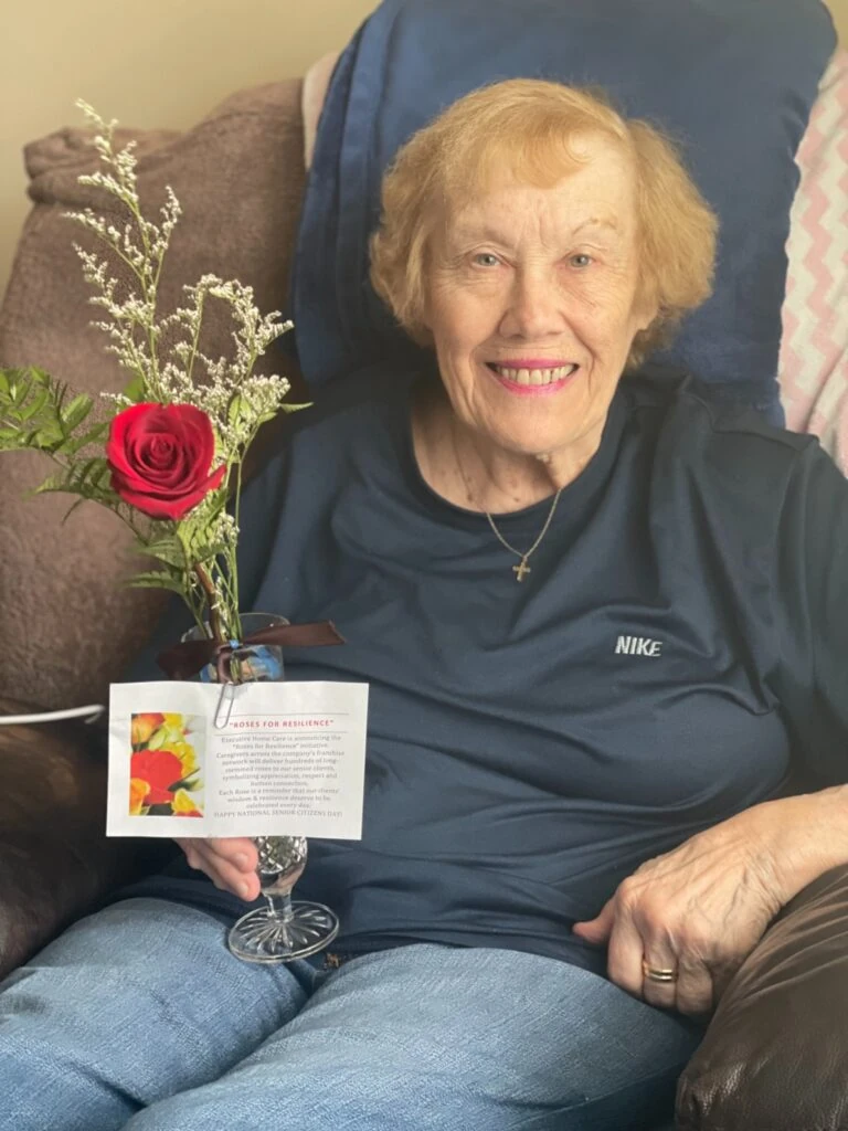 A smiling elderly woman sits in a chair holding a small vase with a single red rose and a “Roses for Resilience” card.