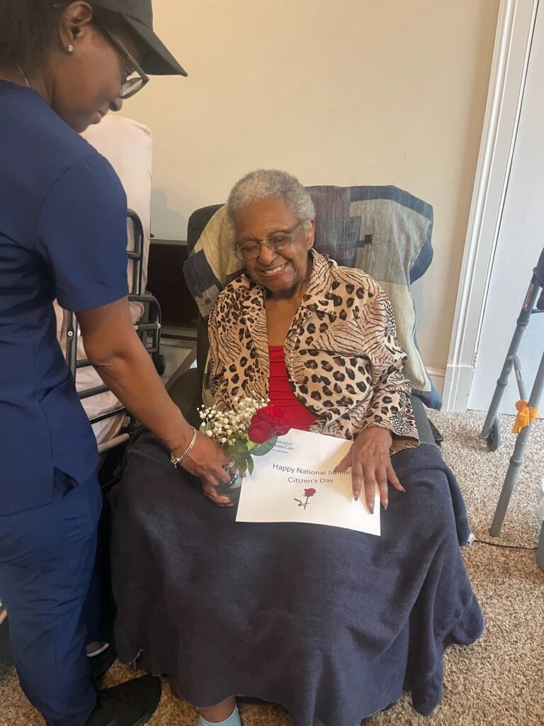 A caregiver places a small vase with a red rose on a blanket as an elderly woman smiles while seated in a chair.