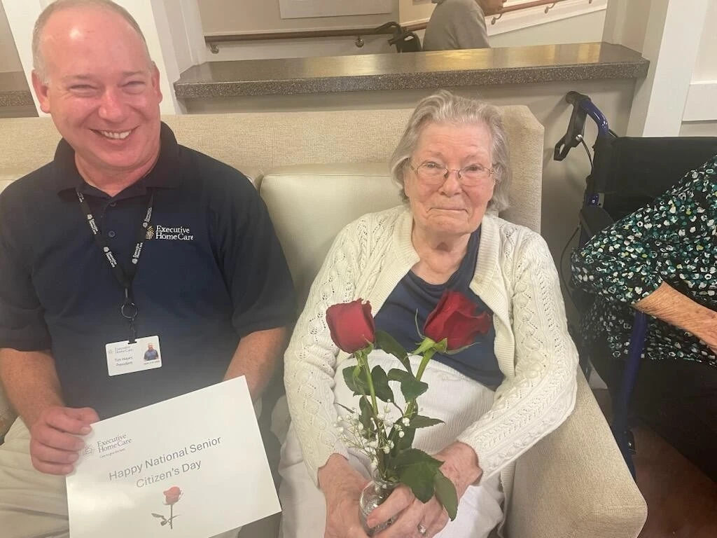 A caregiver sits beside an elderly woman holding two red roses in a vase, with a “Happy National Senior Citizens Day” sign.