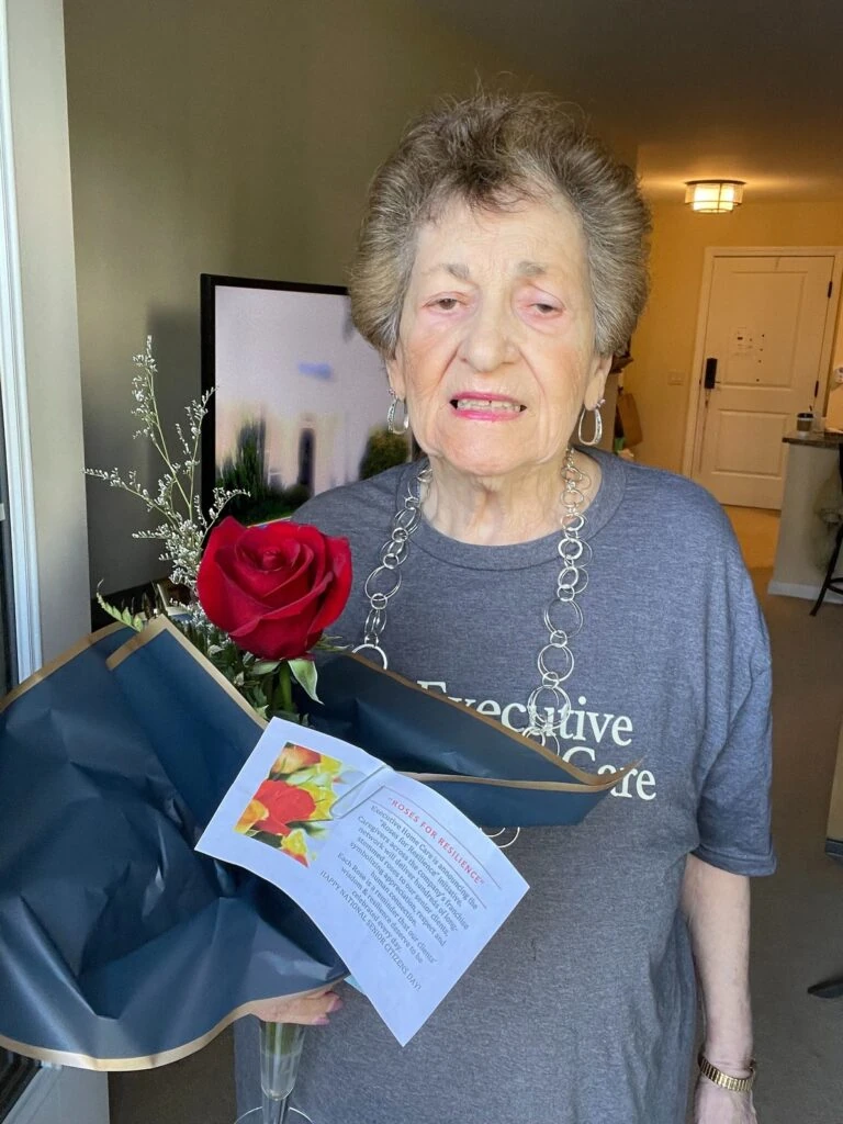 An elderly woman stands indoors holding a wrapped bouquet with a single red rose and a “Roses for Resilience” card.