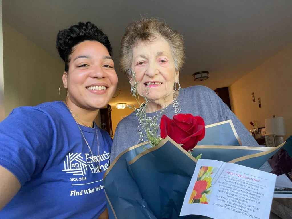 A caregiver and an elderly woman smile together while holding a bouquet with a single red rose and a “Roses for Resilience” card.