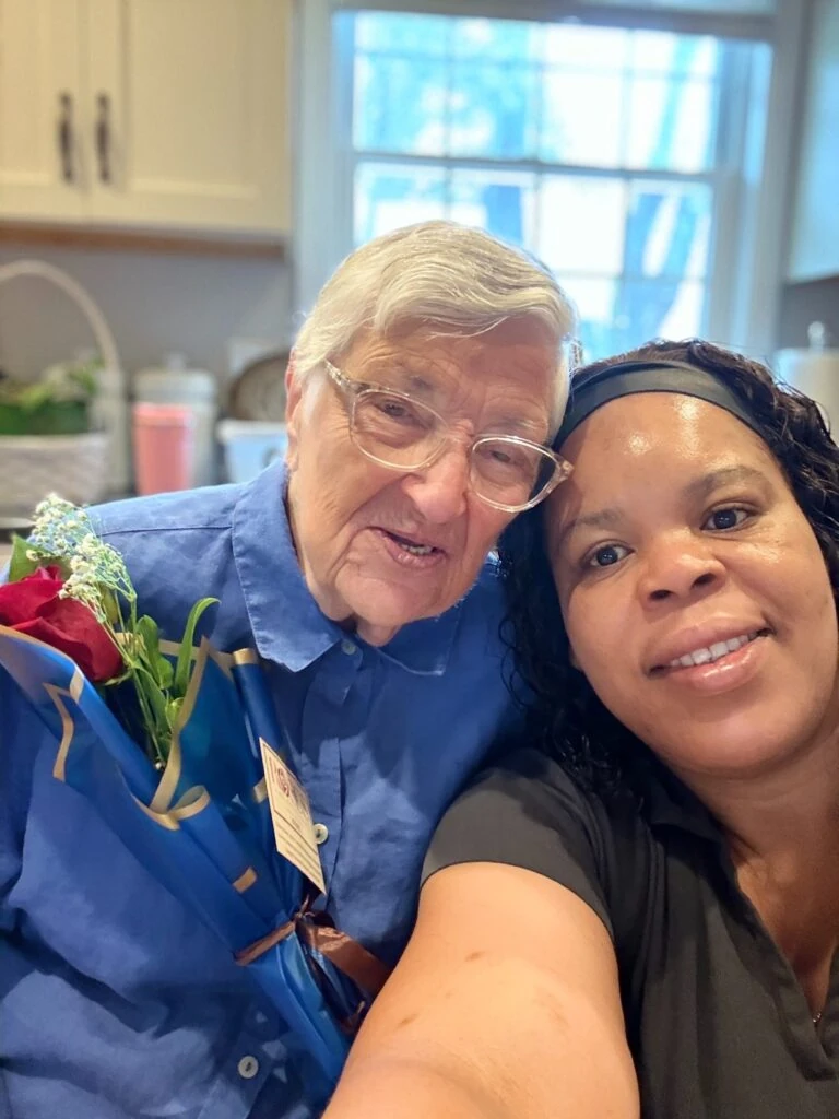 An elderly man and a caregiver take a close selfie together in a kitchen while holding a wrapped red rose.