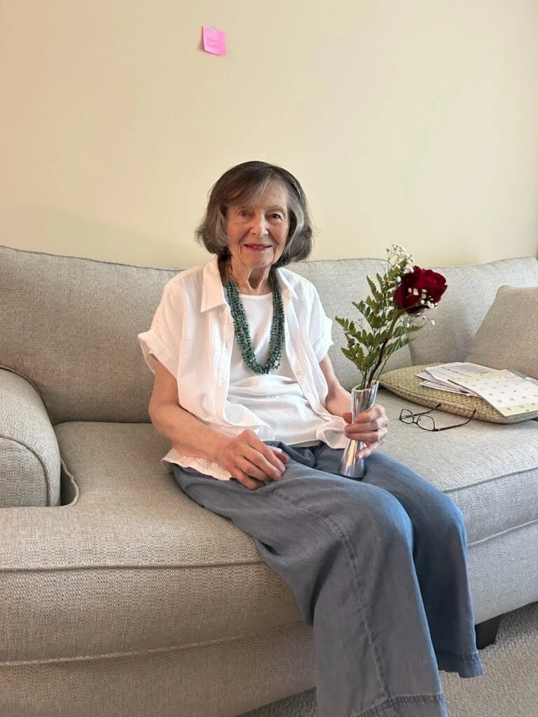 An elderly woman sits on a couch holding a small vase with a single red rose and greenery.