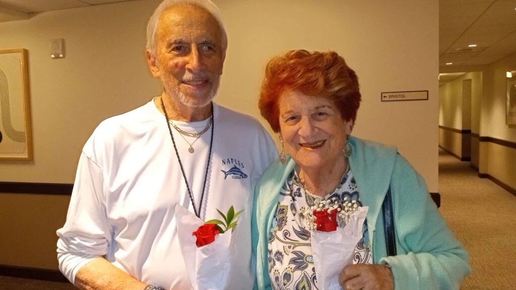 An elderly couple stands in a hallway smiling while each holds a wrapped red rose.